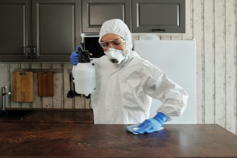 A person in protective clothing disinfects a kitchen counter indoors, focusing on safety and cleanliness.