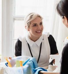 Two housekeepers sharing cleaning supplies, smiling and interacting, indoors.